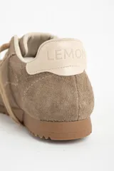 Zapatilla deportiva de gamuza color visón, con cordones a tono y suela de goma marrón. Presenta un diseño retro de running con el nombre de la marca grabado en dorado en el lateral.