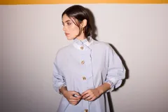 Camisa celeste a rayas verticales azules y marrones, con cuello blanco, cierre frontal con botones dorados y volantes verticales.