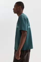 Remera boxy de punto liviano de algodón en color verde oscuro, de hombros caídos, con cuello redondo acanalado y bajo recto. Presenta un diseño estampado en el pecho con texto y logotipo.