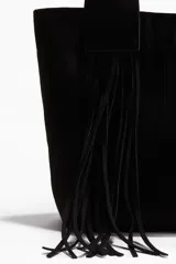 Bolso tipo shopper de color negro, confeccionado en tejido cepillado (similar a gamuza o nobuk). Presenta dos asas anchas con flecos largos colgantes en la parte frontal. Cierre magnético oculto en la parte superior y forro interior de tafeta con bolsillo con cierre.