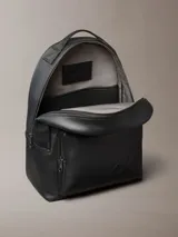 Mochila Calvin Klein de poliuretano color negro, de diseño minimalista con silueta redondeada. Cuenta con un compartimento principal con cierre, bolsillo frontal externo con cierre y el monograma CK grabado. Incluye asa superior y correas acolchadas ajustables.