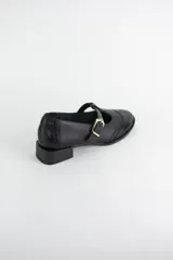 Zapato tipo guillermina estilo oxford, confeccionado en cuero negro con detalles de perforaciones decorativas en la puntera y cierre mediante tira con hebilla metálica dorada. Presenta un taco bajo cuadrado.