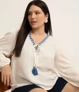 Blusa color blanco con escote en pico con bordado de corazones azules, mangas largas con puños elásticos, lazos con borlas y detalle de hilo metalizado.