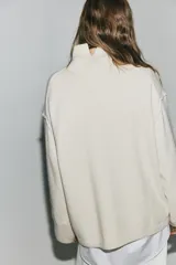 Buzo oversized de punto suave, color beige, con cuello alto acanalado y abertura posterior en V. Presenta hombros caídos, costuras decorativas verticales en el frente y terminaciones acanaladas en puños y bajo.