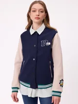 Campera estilo varsity con cuerpo en color marrón y mangas en tono beige. Presenta cierre frontal con botones a presión, cuello, puños y ruedo con franjas tejidas en verde y blanco. Incluye un parche con la letra R en el pecho y detalles bordados en las mangas.