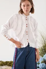 Camisa de lino blanca con cuello mao, manga larga y botones frontales. Presenta volados delanteros con terminaciones desflecadas y detalles de costuras en hilo rojo.