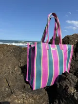 Bolso tipo tote bag con estampado a rayas verticales en tonos verde, rosa, celeste y marrón. Tiene asas de hombro y un parche con la inscripción "AMÉN".