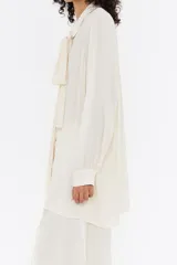 Camisa color crema de corte recto, con cuello camisero con lazada, manga larga y cierre frontal con botonadura.