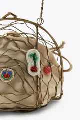 Bolso rígido con estructura metálica dorada y forro interior de tela color beige ajustable con lazada. Decorado con charms colgantes de diferentes motivos y asa superior rígida.