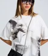 Remera de corte boxy en algodón, color blanco con estampa artística de ángeles en escala de grises. Presenta cuello redondo y mangas cortas.