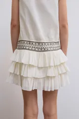 Vestido corto de gasa color blanco, con escote en V cruzado, sin mangas y diseño con volantes en el bajo. Presenta un detalle de tachas plateadas en la cintura.