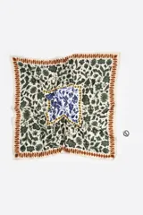 Pañuelo tipo twilly diseñado como charm para bolso, con estampado floral e ikat en tonos azul, verde, amarillo y naranja sobre fondo blanco. Incluye un anillo metálico plateado en forma de corazón que sujeta el pañuelo.