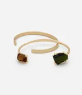 Kit de dos brazaletes abiertos de metal con acabado dorado. Uno de los brazaletes presenta detalles geométricos en sus extremos con incrustaciones de piedras en tonos verde y ámbar.