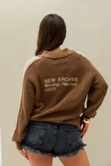 Sweater tejido marrón de manga larga con cuello polo, bandas blancas laterales en las mangas y bordado decorativo en la espalda.