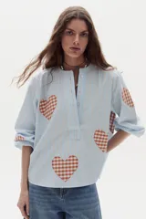 Camisa de algodón celeste a rayas verticales con parches estampados de corazones en tartán marrón y blanco. Posee cuello mao, cierre frontal parcial con botones y mangas largas abullonadas.