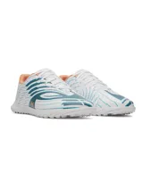 Championes de fútbol Under Armour Shadow Club 4 Turf, diseñados para césped artificial. Presentan un diseño en color blanco con detalles gráficos en azul y toques en naranja. Cuentan con empeine sintético, cordones centrales y suela de goma con tacos multidireccionales para un agarre óptimo.