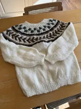 Sweater tejido a mano en lana mohair color crudo, con cuello redondo y mangas largas. Presenta un diseño con motivos geométricos en tonos marrón y verde en la parte superior.