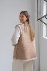 Chaleco de gamuza color beige con interior y detalles de corderito color crudo. Presenta solapas amplias, bolsillos frontales aplicados y diseño sin mangas.