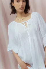 Vestido corto blanco de lino, con diseño suelto y holgado, mangas amplias hasta los codos, escote redondo con abertura en V y lazo decorativo, y un volante en la parte inferior.