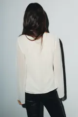 Camisa de georgette color blanco con escote cuadrado, mangas largas acampanadas y forro interior. Presenta detalles de puntilla a tono en el escote y un lazo de terciopelo negro en el centro del pecho.