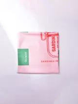 Pareo de tela, color rosa pálido, con estampado gráfico que simula una lata de sardinas en rojo y detalles en verde. Incluye un parche con el texto "Guapa!".