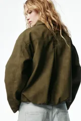 Campera bomber color verde militar de corte oversize, con cuello clásico, cierre frontal oculto bajo solapa y bolsillos laterales ribeteados.
