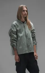Campera rompevientos de diseño oversize, color verde, confeccionada en microfibra técnica ligera. Presenta capucha integrada, cierre frontal técnico y un sistema de frunces elásticos regulables a lo largo de las mangas que permiten ajustar el volumen de la prenda.