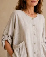 Blusa beige de lino de corte holgado y silueta oversize, con cuello redondo, cierre frontal de botones, bolsillos aplicados grandes y mangas tres cuartos con detalles de perforaciones bordadas en las mangas y el ruedo.
