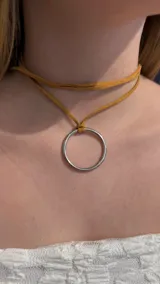 Gargantilla tipo choker compuesta por una tira de gamuza color tostado que se anuda alrededor del cuello, con un dije central en forma de argolla metálica plateada.