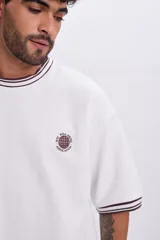Remera manga corta blanca de corte holgado, con cuello redondo y puños acanalados en tono bordó con líneas blancas. Cuenta con un pequeño bordado circular en el pecho con la inscripción "NYC Well-Being Association" y un diseño de red.