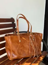 Bolso de cuero marrón con doble asa de hombro, detalles de hebillas metálicas doradas y tiras flecadas colgantes.