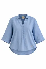 Blusa azul de lino con cuello camisero, escote en V y mangas tres cuartos con vuelta. Presenta un diseño holgado con dobladillo redondeado y un pequeño parche con logo bordado en el lateral inferior.