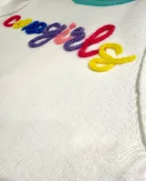 Sweater de punto de manga corta en color blanco, con cuello redondo en color turquesa, puños en colores amarillo y rojo, y borde inferior en color fucsia. Presenta la palabra 'cowgirls' bordada en relieve con letras multicolores.