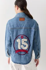 Campera de jean de corte cropped, con cierre frontal de botones metálicos. Presenta un detalle decorativo tejido a mano en crochet con bordes rojos en la solapa y un parche lateral. El diseño incluye bolsillos frontales con solapa y un bordado en la parte posterior, con un corte asimétrico más largo en la espalda.