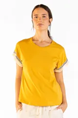 Remera de manga corta color mostaza, confeccionada en 100% algodón, con cuello redondo y detalles de flecos y guardas geométricas en las mangas.