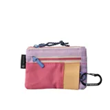 Billetera tipo pouch con múltiples compartimentos y cierres. Diseño color block en tonos pastel y vibrantes: lila, rosa, beige y detalles en azul. Incluye un mosquetón negro con el logo de la marca y un tirador de cierre azul claro.