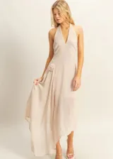 Vestido largo de tela fluida en color beige, con escote halter y detalle de flor aplicada en la cintura. Presenta un diseño asimétrico con caída suave y elegante.
