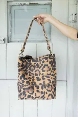 Cartera tipo bolso de hombro con diseño de estampado animal print de leopardo. Cuenta con una correa fija de longitud media y herrajes metálicos en tono bronce envejecido que conectan la correa con el cuerpo del bolso.
