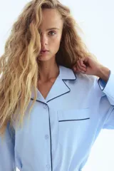 Camisa oversized de algodón color celeste claro con ribetes en contraste negro. Presenta cuello abierto, cierre frontal de botones, un bolsillo superior de parche y mangas largas con puños anchos.