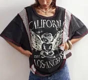 Remera de algodón color negro con efecto lavado, corte oversize y mangas cortas. Presenta un estampado gráfico frontal de un ángel con la inscripción 'California Los Angeles' y detalles de encaje en los laterales con costuras a contratono.