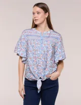 Blusa holgada de poliéster con estampado floral y geométrico en tonos celestes, azules y anaranjados sobre base blanca. Presenta cuello a la base con escote en V y botones frontales, mangas cortas amplias de estilo mariposa y un nudo en el ruedo.