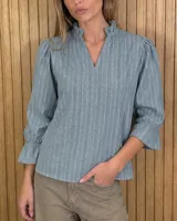 Blusa color lila con estampado vertical de ramas y hojas blancas, cuello alto con volados y escote en V. Presenta mangas largas abullonadas con puños elastizados.