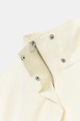 Blazer corto de color marfil con cuello subido, manga larga y bolsillos laterales. Cuenta con un cinturón del mismo tejido para anudar en la cintura, forro interior a tono y cierre frontal con cremallera y botones a presión ocultos bajo solapa.