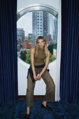 Top sin mangas color verde oliva, con cuello alto y cierre frontal de botones metálicos decorativos. Presenta un corte ajustado en el torso que se ensancha ligeramente hacia el bajo, con detalles de trabillas horizontales a la altura de la cadera.