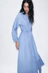 Vestido midi de corte camisero con estampado de rayas verticales en tonos azul, blanco y rojo. Presenta cuello redondo con escote en pico, mangas largas abullonadas con puños elásticos y cinturón ajustable del mismo tejido. Incluye bolsillos laterales ocultos y cierre frontal cruzado con botones.