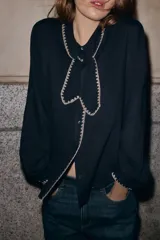 Blusa de color negro con cuello solapa, lazo frontal y manga larga. Presenta detalles de bordados en color crema en los bordes del cuello, el lazo, la tapeta de botones y los puños. Cierre frontal con botones.