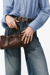 Cartera bandolera rectangular de color marrón, con diseño alargado y asas cortas de mano. Incluye un detalle de tira decorativa con hebillas metálicas en la parte frontal y herrajes dorados.