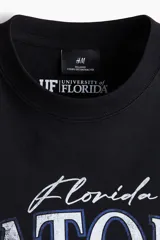Remera de algodón negro con hombros caídos y cuello redondo. Estampado frontal estilo universitario con la inscripción 'Gators', casco de fútbol americano e insignias conmemorativas.