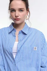 Camisa oversize de popelina a rayas celestes y blancas, con cuello camisero, manga larga con puños abotonados, cierre frontal con botones y un bolsillo en el pecho con etiqueta bordada.