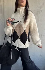 Sweater de punto grueso con cuello alto, diseño de rombos en tonos beige y negro sobre base color natural, de corte holgado y mangas largas con puños ajustados.